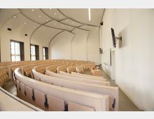 Ein moderner Hörsaal mit halbrunden, ansteigenden Sitzreihen aus hellem Holz, mehreren großen Fenstern an der linken Wand, einer weißen Tafel und einem Rednerpult vorne sowie Wandmonitoren an der rechten Wand. Die gewölbte Decke ist mit eingebauten Leuchten ausgestattet.
