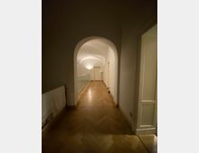 Ein heller Korridor mit einem hölzernen Fischgrätboden und weiß gestrichenen Wänden, der durch eine gewölbte Decke definiert wird. Links befindet sich ein Handtuchhalter, rechts eine Glastür, und am Ende des Flurs steht eine geschlossene weiße Tür.