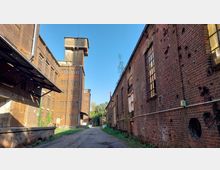 Industriegelände mit Backsteingebäuden, darunter ein hohes Turmgebäude mit kleinen rechteckigen Fenstern. Eine schmale Straße mit Schienen läuft zwischen den Gebäuden entlang, umgeben von vereinzeltem Grün und blauer Himmel im Hintergrund.