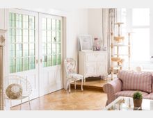 Heller Wohnraum mit Holzboden, weißen Schiebetüren mit grün eingerahmtem Milchglas, dekorativen Möbeln im Vintage-Stil und einer Katzenecke mit einem Kratzbaum. Auf einem Beistelltisch steht eine kleine Pflanze vor einem gestreiften Sessel.