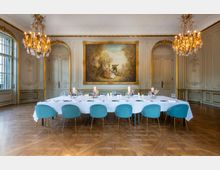 Eleganter Speisesaal mit einer langen, weiß gedeckten Tafel und türkisfarbenen Stühlen, umgeben von reich verzierten Wänden mit goldenen Akzenten, zwei Kronleuchtern und einem großen barocken Gemälde in der Mitte der Wand. Der Raum hat einen Holzboden und bogenförmige Fenstertüren.