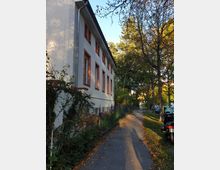 Seitenansicht eines zweistöckigen Gebäudes mit weißer Fassade und roten Fensterrahmen, flankiert von einem Gehweg mit Herbstlaub. Im Hintergrund stehen Bäume, und rechts neben dem Gehweg sind geparkte Autos zu sehen.
