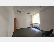 Ein leerer Raum mit weiß gestrichenen Wänden, grauem Teppichboden und einer abgehängten Rasterdecke. Der Raum enthält ein kleines Waschbecken mit Wandfliesen, einen Heizkörper unter einem Fenster mit vertikalen Jalousien sowie einige blaue Stühle und einen schmalen Holzschrank.