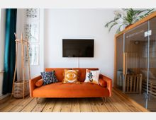 Wohnzimmer mit weißer Wand, einem orangenen Sofa mit gemusterten Kissen, einem an der Wand befestigten Flachbildfernseher, einem Holz-Kleiderständer und einer kleinen Heimsauna mit Glastür. Holzboden und Pflanzen verleihen dem Raum eine einladende Atmosphäre.
