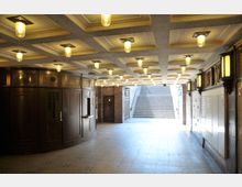 Unterirdischer Durchgang mit dunklem Holzpavillon auf der linken Seite, einer kassettierten Decke mit warm leuchtenden Lampen und einer Treppe, die ins Freie führt. Die Wände sind mit dekorativen Elementen und Fliesen gestaltet.