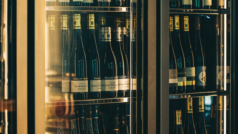 Gläserner Weinkühlschrank mit Metallrahmen und mehreren Regalböden, dicht bestückt mit Wein- und Sektflaschen. Warmes Licht und Spiegelungen in den Glastüren lassen ein Innenraum‑Ambiente wie in einer Bar oder einem Restaurant erkennen.