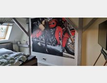 Schlafzimmer in einem Dachgeschoss mit sichtbaren Holzbalken, einem Bett links und einem großen Wandbild, das einen Teil eines roten Motorrads und eine Person zeigt. Ein Dachfenster sorgt für Tageslicht.