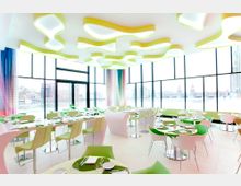 Heller, moderner Restaurantbereich mit großen, bodentiefen Fenstern, die einen Blick auf einen Fluss und städtische Gebäude im Hintergrund bieten. Der Raum ist mit grünen und weißen Möbeln sowie geschwungenen Deckenleuchten gestaltet.