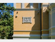 Seitenfassade eines gelblichen Ziegelgebäudes mit hellen Gesimsen und einer dekorativen Reliefplatte in weißem Rahmen. Links ragen hohe Bäume und Grün in das Bild, rechts sind Gebäudeecken und Fallrohre zu sehen.