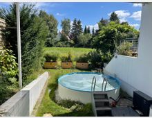 Garten mit kleinem runden Pool, umgeben von Rasen und Pflanzen. Im Hintergrund befinden sich Hochbeete, eine Hecke und eine freie Wiese mit vereinzelten Bäumen.