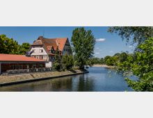 Fachwerkgebäude mit roten Ziegeldächern am Ufer eines Flusses, umgeben von Bäumen und Grünflächen unter einem klaren, blauen Himmel. Im Vordergrund sind ein Wasserlauf und eine gepflasterte Uferpromenade sichtbar.