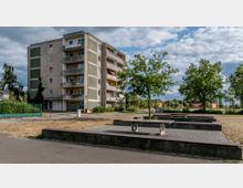 Wohnhaus mit fünf Stockwerken in einfacher Bauweise, umgeben von Bäumen und Grünflächen. Im Vordergrund befindet sich ein gepflasterter Platz mit Sitzbänken und Tischkonstruktionen aus Beton.