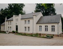 Weiße, zweigeschossige Villa im Landhausstil mit grauem Schieferdach, symmetrischer Fassade und zwei runden Türmchen am Eingangsbereich. Davor liegen ein Kieshof und ein schmaler bepflanzter Gartenstreifen, umgeben von hohen Bäumen.