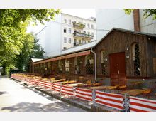 Holzgebäude mit einer langen Terrasse, die von rot-weißen Absperrbändern umgeben ist. Vor dem Gebäude stehen zahlreiche Tische und Stühle unter einem gestreiften Sonnendach, im Hintergrund sind Bäume und ein Wohnhaus mit Balkonen zu sehen.