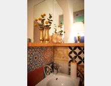 Waschbecken in einem kleinen Badezimmer mit farbenfrohen, gemusterten Fliesen an der Wand. Auf einer Holzablage über dem Waschbecken stehen goldene Vasen mit gelben Blumen und ein Spiegel reflektiert die Umgebung.
