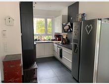 Moderne Küche mit einer Kombination aus weißen und schwarzen Schränken, schwarzer Arbeitsplatte und Edelstahlgeräten. Ein großes Fenster bietet Blick ins Grüne, und auf der linken Seite steht ein rotes Metallschränkchen neben einer schwarzen hölzernen Sitzbank.