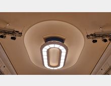 Ansicht einer Decke mit elliptischem Deckenleuchter in einem historischen Saal. Der Leuchter ist von verzierten Elementen umgeben, und Beleuchtungstraversen mit Scheinwerfern sind an den Seiten montiert.