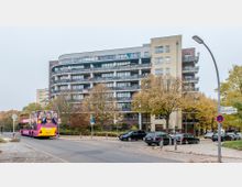 Mehrstöckiges Wohngebäude mit modernen Balkonen, umgeben von herbstlich gefärbten Bäumen und parkenden Autos. Im Vordergrund fährt ein gelb-pinker Doppeldeckerbus eine Straße entlang, Straßenlaternen und ein Straßenschild sind sichtbar.