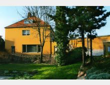 Zweistöckiges Wohnhaus mit gelber Fassade und rotem Satteldach, umgeben von Bäumen und einem Garten mit grüner Rasenfläche und dichten Sträuchern. Im Vordergrund stehen hohe Bäume und Pflanzenbeete, die teilweise von Sonnenlicht beleuchtet werden.