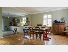 Helles Esszimmer mit Holzdielenboden, einem großen Holztisch und verschiedenfarbigen Stühlen; im Hintergrund eine offene Küche mit weißen Schränken, offenen Regalen und einer Arbeitsfläche. Zwei Fenster lassen Tageslicht herein, rechts ein hölzernes Buffet mit blauen Vasen und einer Lampe.