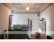 Rustikale Küche mit unverputzter, teils beschädigter Decke, grün gestrichener Kochinsel, Edelstahlkühlschrank, Holzesstisch mit Obstschale und weißem Wandschrank mit Globus darauf. Die Wände sind schlicht weiß gehalten, mit minimaler Dekoration.