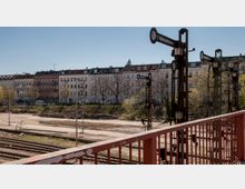 Ehemaliger Bahnhofsbereich mit mehreren alten Stellwerksignalen im Vordergrund, umgeben von Bahngleisen und einer Freifläche, im Hintergrund eine Reihe von Wohngebäuden im klassischen europäischen Baustil.