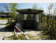 Überdachter Eingang zu einer Unterführung mit Glaswänden, umgeben von Bäumen und Sträuchern. Im Hintergrund sind ein Gleisbereich mit abgestellten Zügen sowie ein Fahrrad am rechten Bildrand zu sehen.