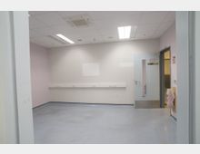 Leerer Raum mit weißen Wänden und einem graublauen Boden, beleuchtet durch Deckenleuchten und ein kleines Fenster in der Tür. An den Wänden befinden sich Steckdosenleisten, und neben der Tür hängt ein Feuerlöscher in einer Plastikabdeckung.