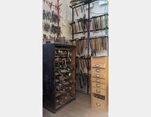 Werkstatt mit verschiedenartigen Werkzeugen. An den Wänden hängen Zangen, Hämmer und andere Handwerkzeuge, daneben ein schwarzer Schrank mit Metallformen und eine Holzkommode mit Schubladen.
