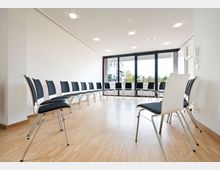 Heller Seminarraum mit einer kreisförmig angeordneten Stuhlreihe, Holzboden, weißen Wänden und einer Fensterfront, die auf eine Außenterrasse mit Blick auf moderne Gebäude führt.