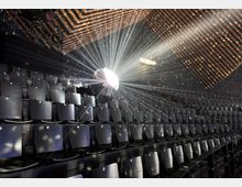 Ein moderner Zuschauerraum mit schwarzen Sitzreihen in ansteigender Anordnung. Eine Disco-Kugel in der Mitte des Raumes reflektiert Lichtstrahlen, die den dunklen Raum und die Holzlamellen an der Decke illuminieren.