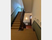 Treppenhaus in einem Altbau mit Teppichboden auf den Stufen, einem hölzernen Geländer und verzierten Wanddekorationen im Jugendstil. Der Bereich ist durch eine Wandlampe beleuchtet, und eine Holztür mit Fenster befindet sich am unteren Ende der Treppe.