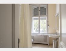 Badezimmer mit einem großen Fenster mit Rollläden und Blick ins Grüne, Waschbecken mit Spiegel, chromfarbenem Mülleimer und einer hellen Duschvorhangtrennwand. Die Wände sind beige, der Boden ist gefliest.