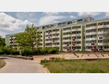 Mehrstöckiges Wohngebäude mit hellgrüner Fassade und Balkonen, umgeben von Bäumen und Büschen. Im Vordergrund befindet sich ein Spielplatz mit einer Rutsche und einem Klettergerüst auf einer Sandfläche.