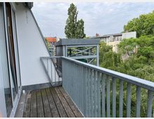Ein Balkon mit Holzfußboden und Metallgeländer an einem modernen Wohngebäude. Im Hintergrund sind Bäume und angrenzende Wohnhäuser erkennbar, unter einem bewölkten Himmel.
