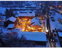 Luftaufnahme eines verschneiten Dorfes bei Dämmerung mit beleuchteten Gebäuden. In der Mitte befindet sich ein Innenhof mit einem geschmückten Weihnachtsbaum, umgeben von alten Backstein- und Fachwerkhäusern.