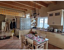 Ländliche Küche mit cremefarbenen Einbauschränken, einer Arbeitsplatte aus Holz und hängendem Kronleuchter. Der Raum hat eine Glassteinwand, freiliegende Holzbalken an der Decke, Fliesenboden und ein Esstisch mit Blumenarrangement.