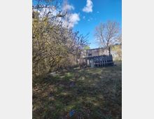 Böschungsfläche mit Gras und niedrigen Sträuchern vor einem kleinen, teils eingegrabenen Betongebäude mit Fensteröffnung. Rechts steht ein dunkles Holzgeländer, im Hintergrund sind Bäume und ein Gewächshaus unter blauem Himmel mit Wolken zu sehen.