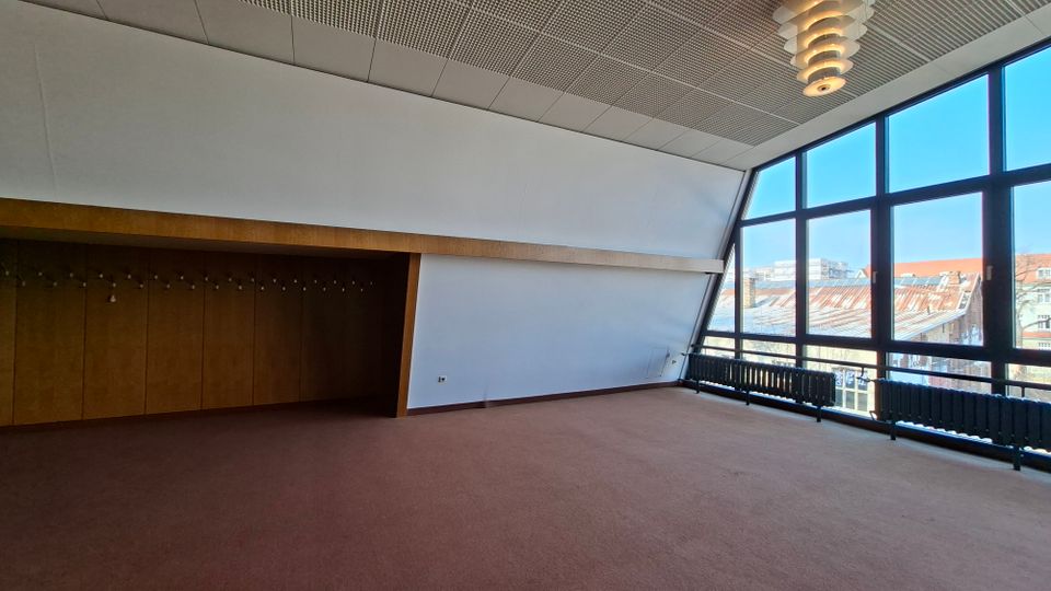 Leerer Innenraum eines modernen Gebäudes mit schräger Decke, braunem Teppichboden und Holzverkleidung an der linken Wand. Rechts sorgt eine große, bodentiefe Fensterfront mit schwarzen Rahmen und Heizkörpern darunter für Tageslicht und Blick auf umliegende Dächer.