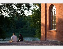 Eine Backsteinfassade eines Gebäudes mit einem großen, gewölbten Fenster auf der rechten Seite, daneben zwei Personen, die auf einer gepflasterten Fläche am Ufer eines Sees sitzen, umgeben von Bäumen.