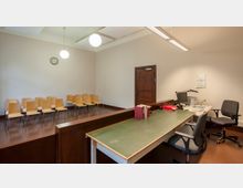 Ein einfacher Gerichtssaal mit einem erhöhten Bereich für Zuschauer mit Holzstühlen, einer Holztür an der Wand und einem Schreibtisch mit Bürostühlen, Computer und Akten im Vordergrund. Zwei Hängelampen und eine Wanduhr sind an der hellen, schlichten Decke angebracht.