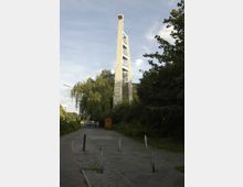 Ein moderner Glockenturm aus hellem Beton mit mehreren offenen Ebenen ragt neben einem Gehweg empor. Die Umgebung ist von Bäumen und Sträuchern geprägt, mit einem asphaltierten Weg und vereinzelten Pollern im Vordergrund.