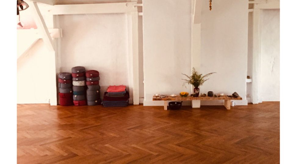 Ein heller Raum mit weiß gestrichenen Holzbalken an der Decke, einem Boden in Fischgrätparkett, einem Kronleuchter und einer schlichten Dekoration. Links stehen gestapelte Yogamatten und Kissen, rechts befindet sich ein niedriger Holztisch mit Pflanzen und Kerzen.