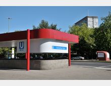 U-Bahnhof-Eingang mit moderner Überdachung in Rot und Weiß, beschriftet mit "Fehrbelliner Platz". Im Hintergrund sind Bäume, Autos und ein mehrstöckiges Gebäude sichtbar.