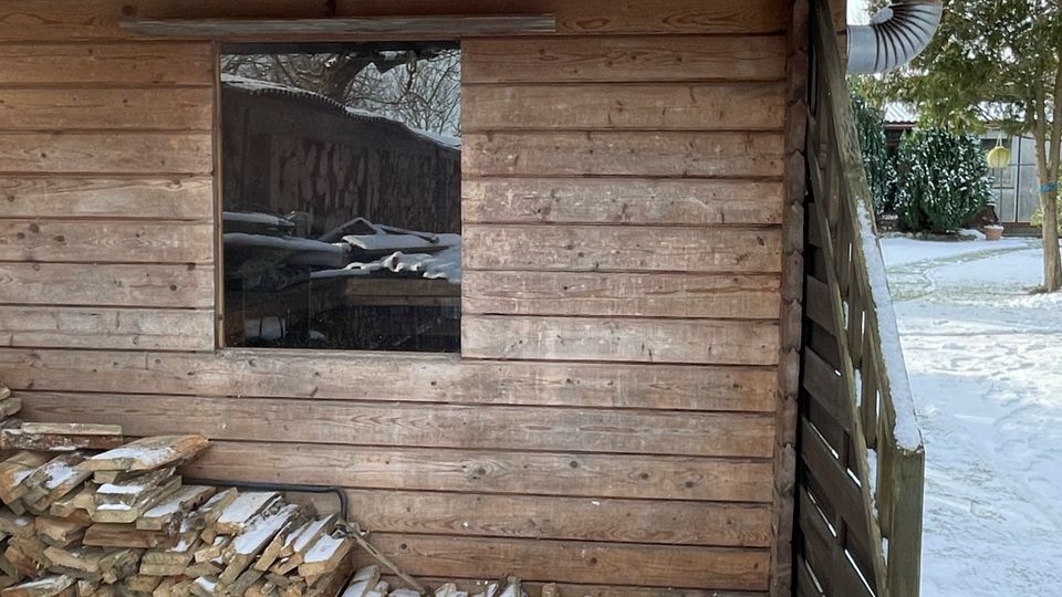 Holzhütte im winterlichen Garten mit schneebedecktem Flachdach, kleinem Fenster und einem Metallkaminrohr an der rechten Seite. Vor der Wand ist ein Stapel Brennholz gelagert, daneben ein Lattenzaun; der Boden und die Umgebung sind verschneit.