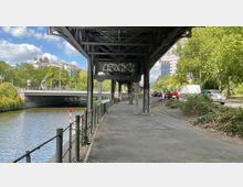 Fußgängerweg unter einer metallenen Hochbahntrasse entlang eines Flusses, daneben eine Straße mit Autos; im Hintergrund Bäume, eine Brücke und moderne Gebäude.
