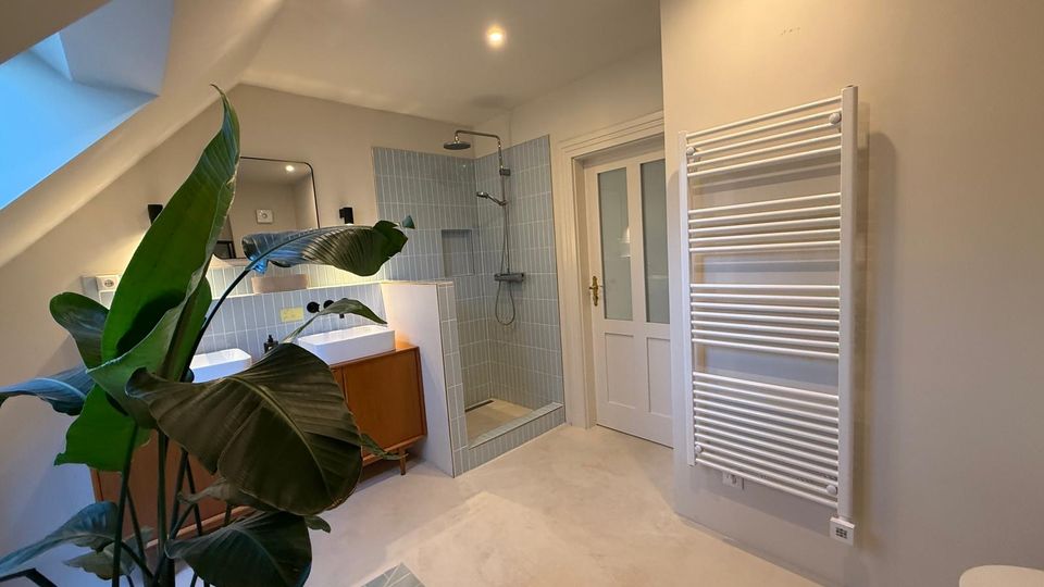 Modernes Badezimmer mit einer offenen Dusche, zwei weißen Waschbecken auf einem Holzunterschrank und einem Handtuchheizkörper. Der Raum ist hell gestaltet mit hellgrauen Fliesen, schräger Decke und einem großen Pflanzenarrangement.