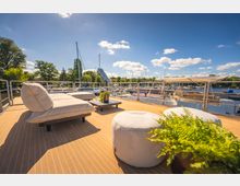 Sonnendeck eines Bootes mit hellen Polstermöbeln und Pflanzen im Vordergrund, im Hintergrund ein Yachthafen mit zahlreichen Segel- und Motorbooten sowie umgebender grüner Vegetation unter klarem, blauem Himmel.
