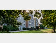 Mehrstöckige Wohngebäude mit Balkonen und schlichter Fassade, umgeben von einer begrünten Landschaft mit Bäumen, gepflegtem Rasen und Gehwegen. Im Vordergrund befinden sich schattenspendende Bäume und Freizeitflächen.