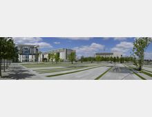 Weitläufiger Platz mit modernen Gebäuden im Hintergrund, darunter das Bundeskanzleramt mit markanter Glasfassade und weißen Betonstreben. Der Platz ist von schmalen Grünstreifen und gepflasterten Wegen durchzogen, umgeben von Bäumen und einem klaren Himmel mit lockeren Wolken.